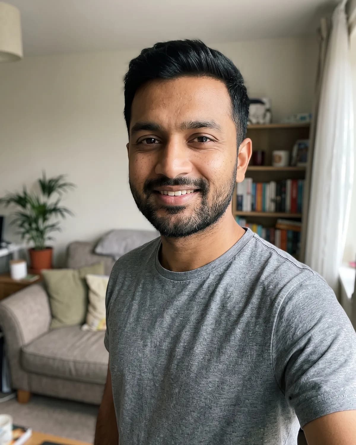 South Asian man with short black hair and a short beard in a grey t-shirt, taking a selfie in a living room with a sofa and bookshelf.