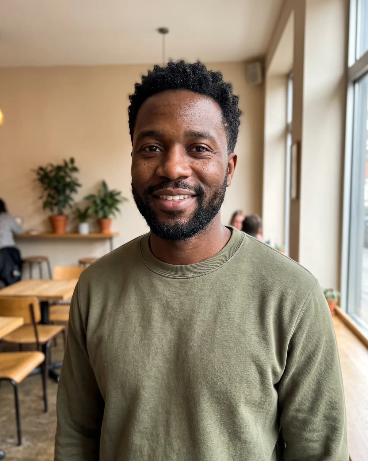 Black man with short curly hair and a neat beard in an olive sweatshirt, smiling in a café.