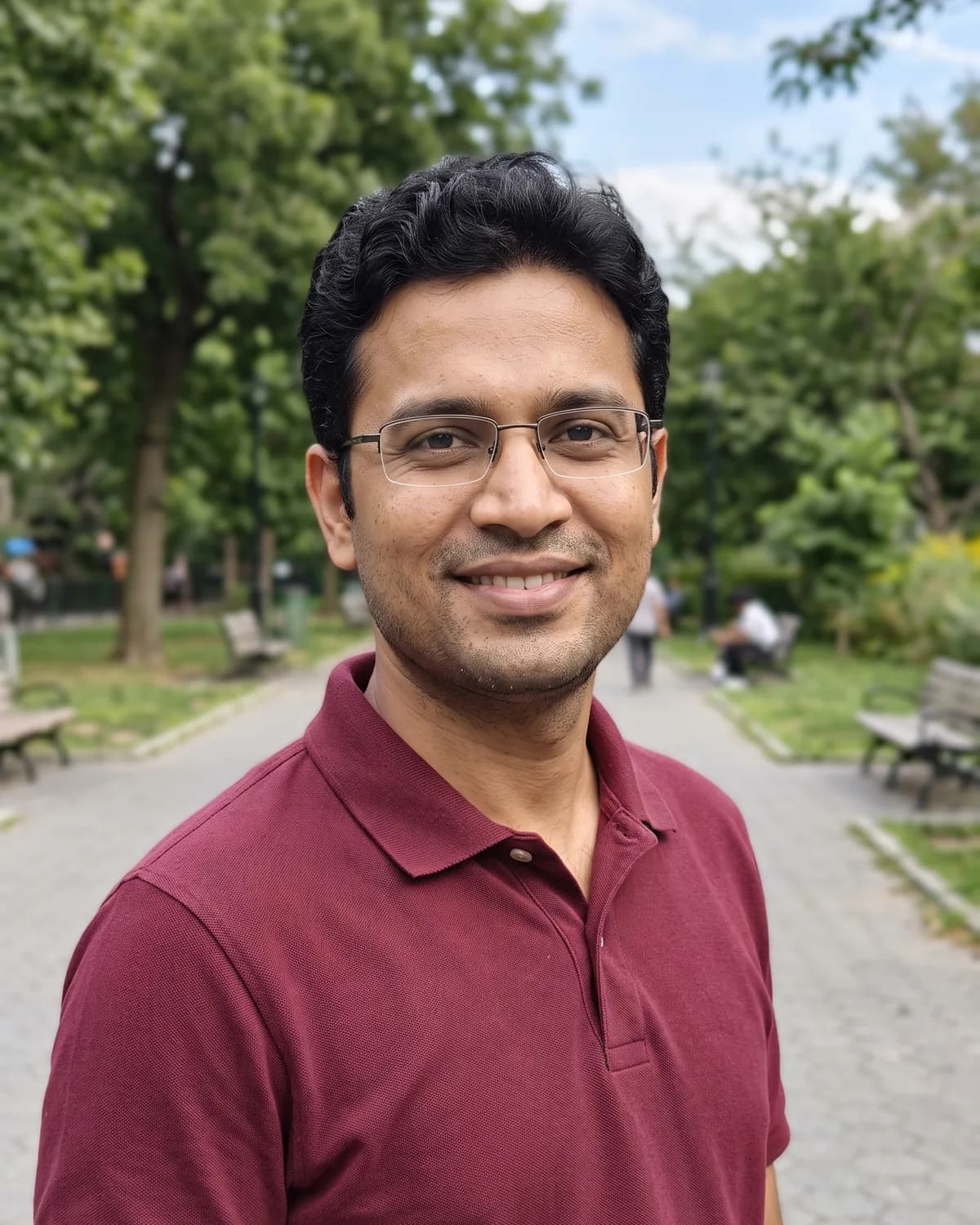 South Asian man with wavy black hair, thin rimless glasses and light stubble in a maroon polo, smiling in a park.