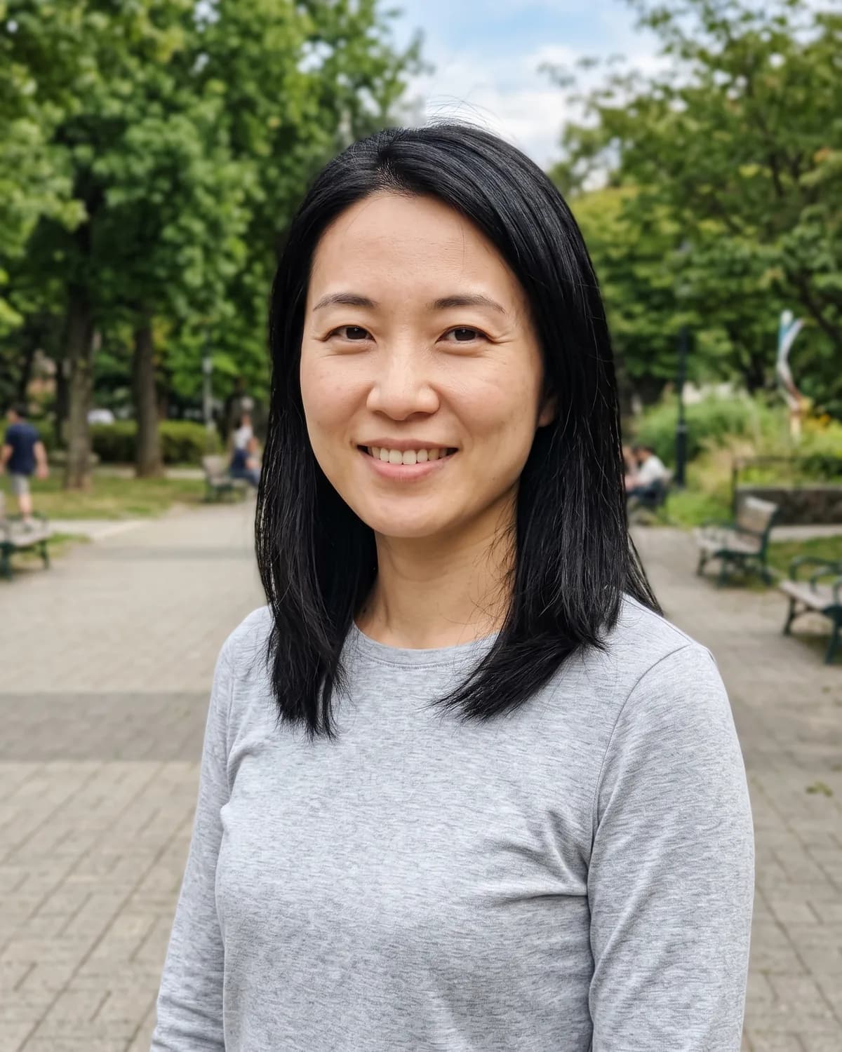 East Asian woman with shoulder-length straight black hair in a grey long-sleeve top, smiling in a park.