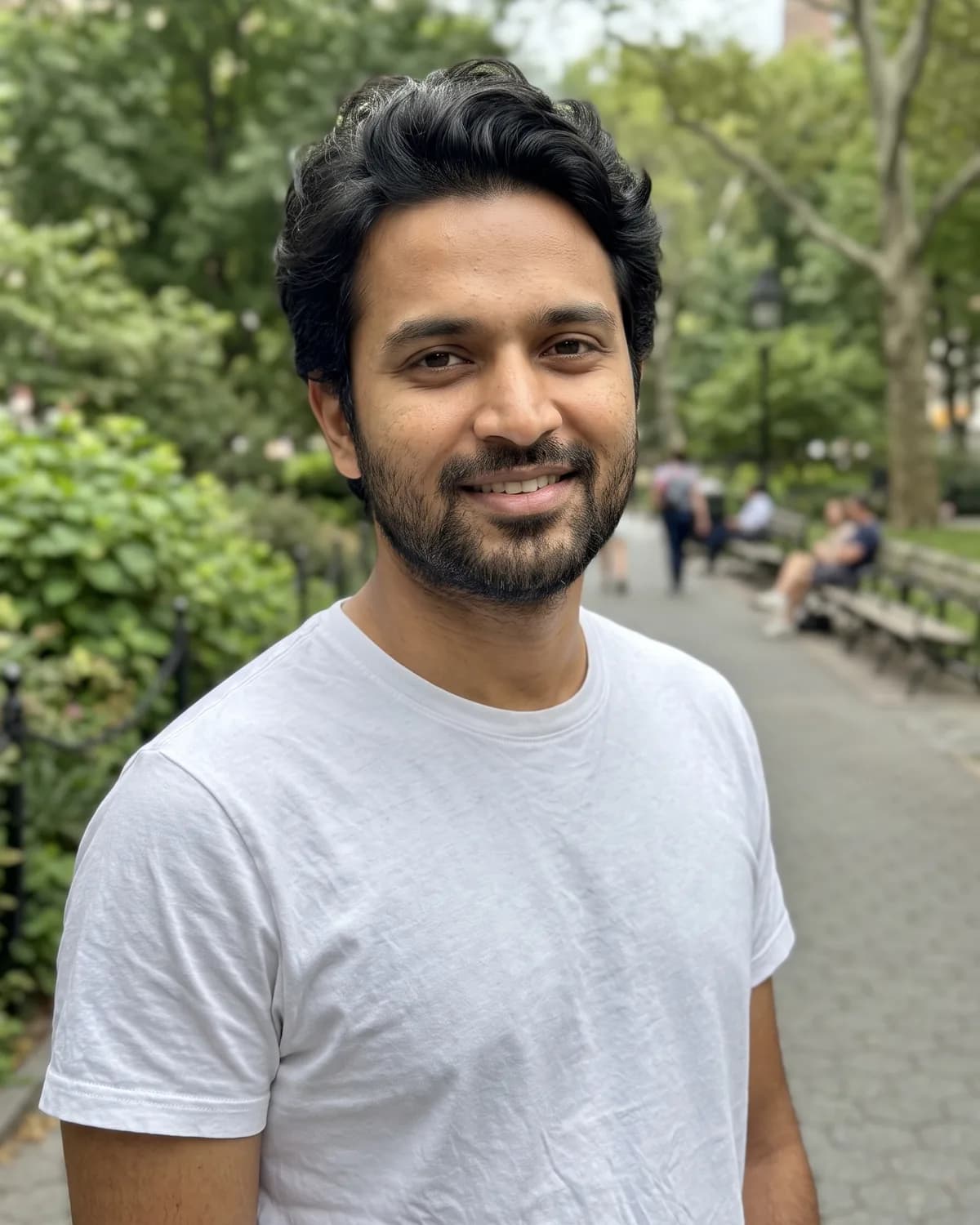 Young South Asian man with wavy dark hair and a beard in a white t-shirt, smiling in a leafy park.