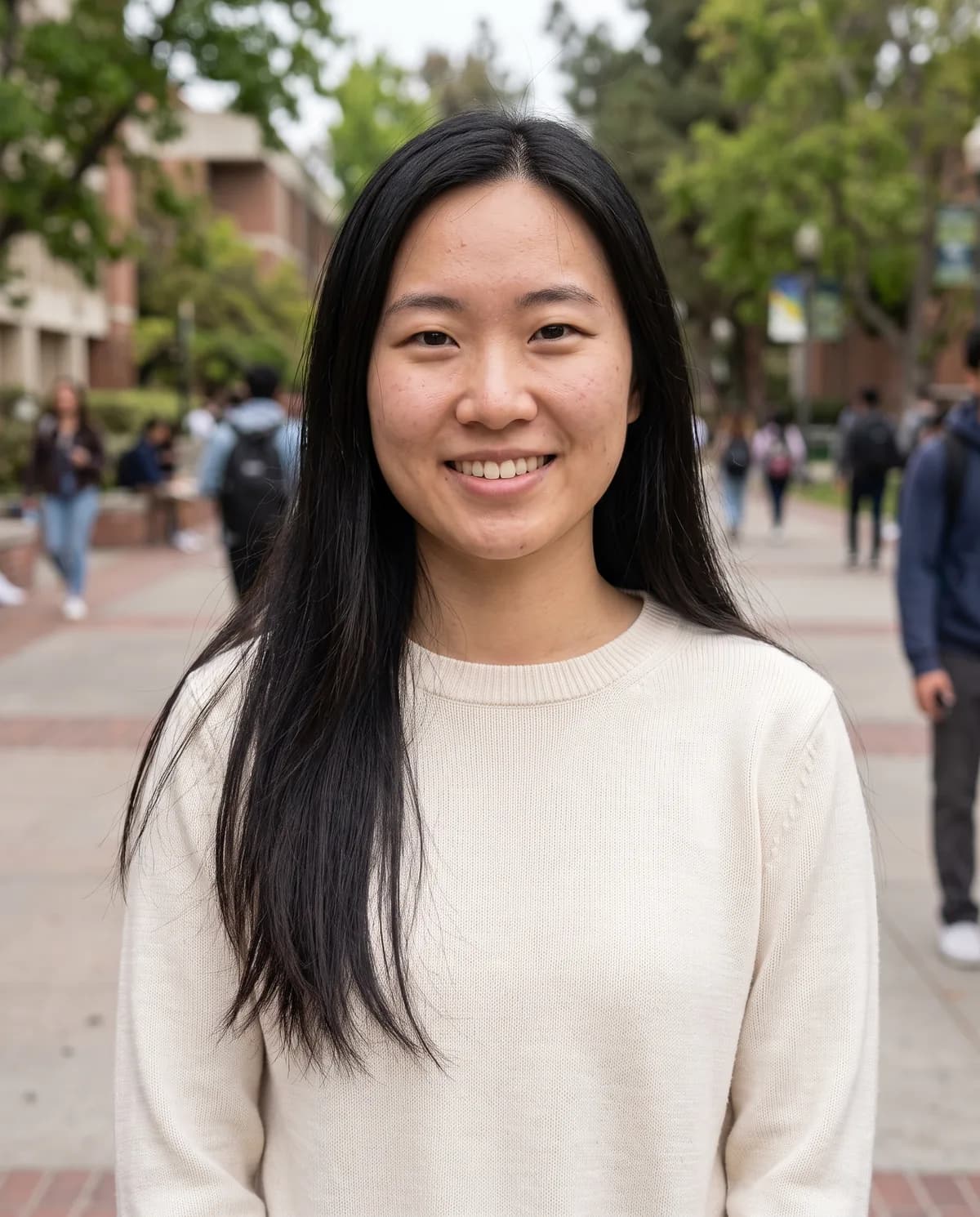 Young East Asian woman with long straight black hair in a cream sweater, smiling on a campus plaza.