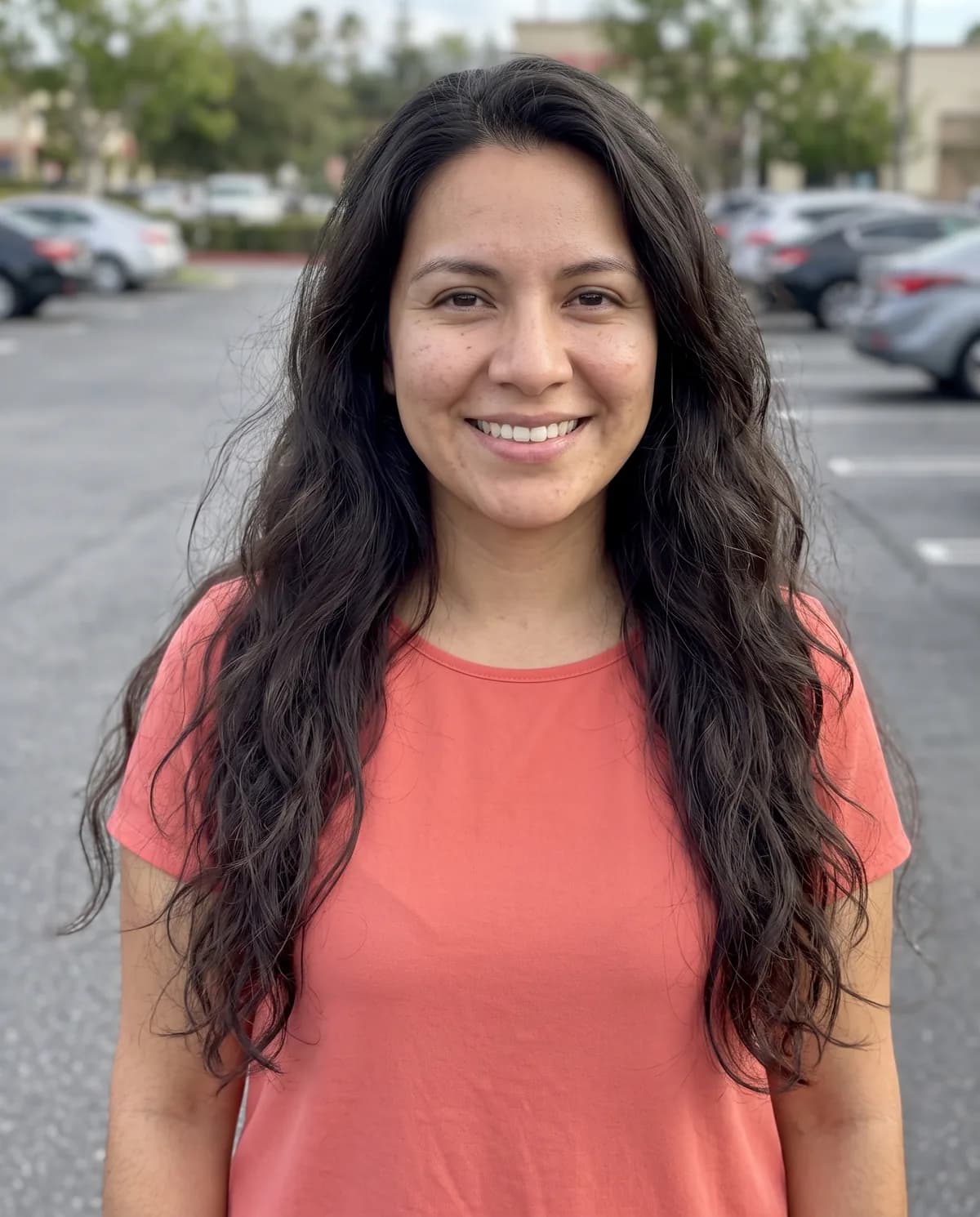 Woman with long wavy dark hair and freckles in a coral t-shirt, smiling in a parking lot.