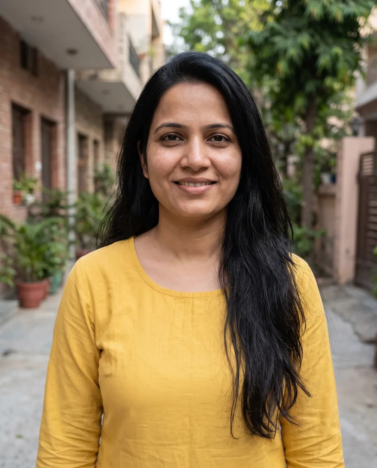 South Asian woman with long wavy black hair in a yellow long-sleeve top, smiling in a residential alley.