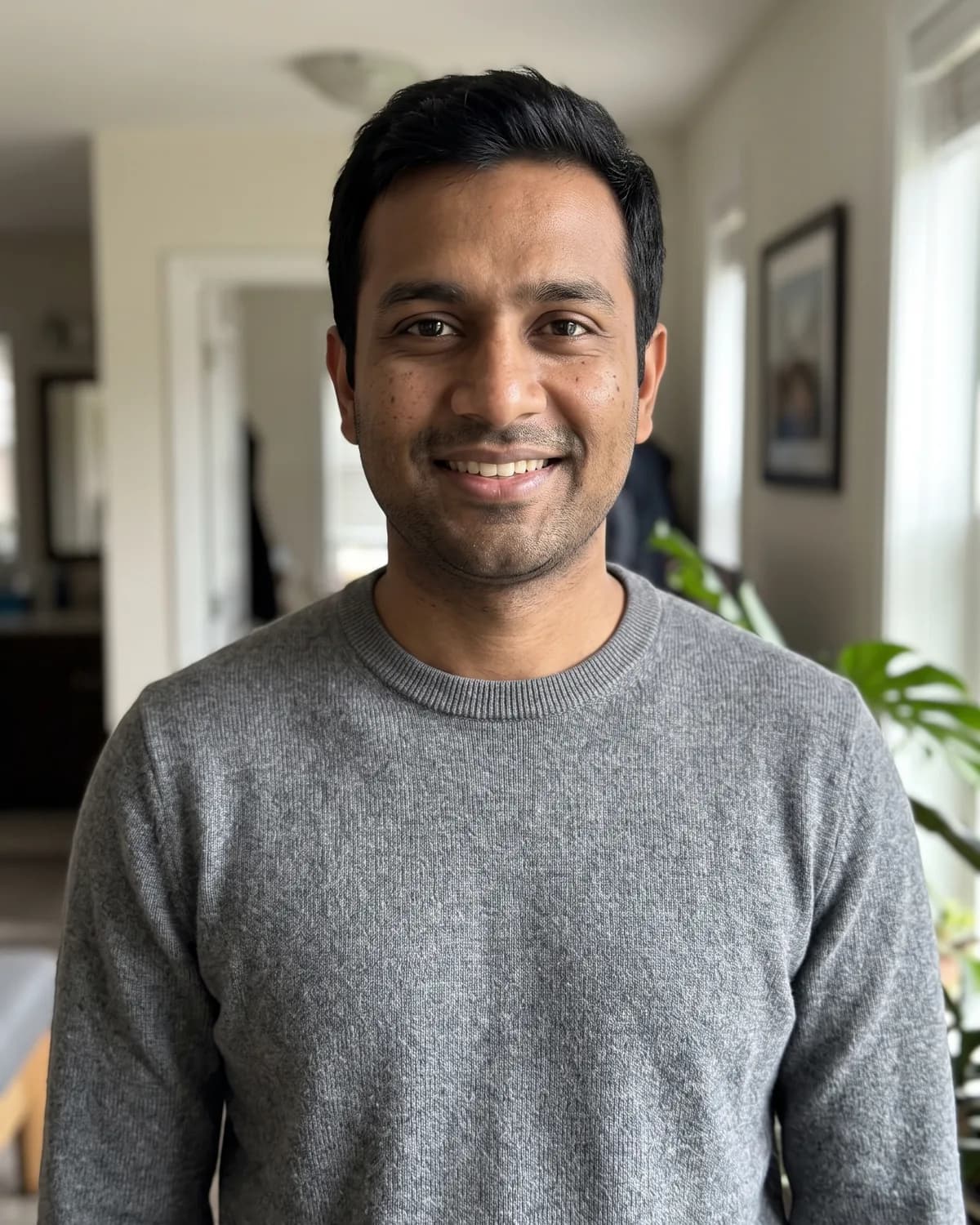 South Asian man with short dark hair and light stubble in a heather-grey sweater, smiling indoors.