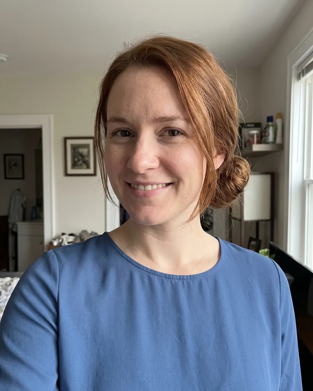 Woman with reddish-brown hair in a messy low bun in a blue blouse, smiling indoors.