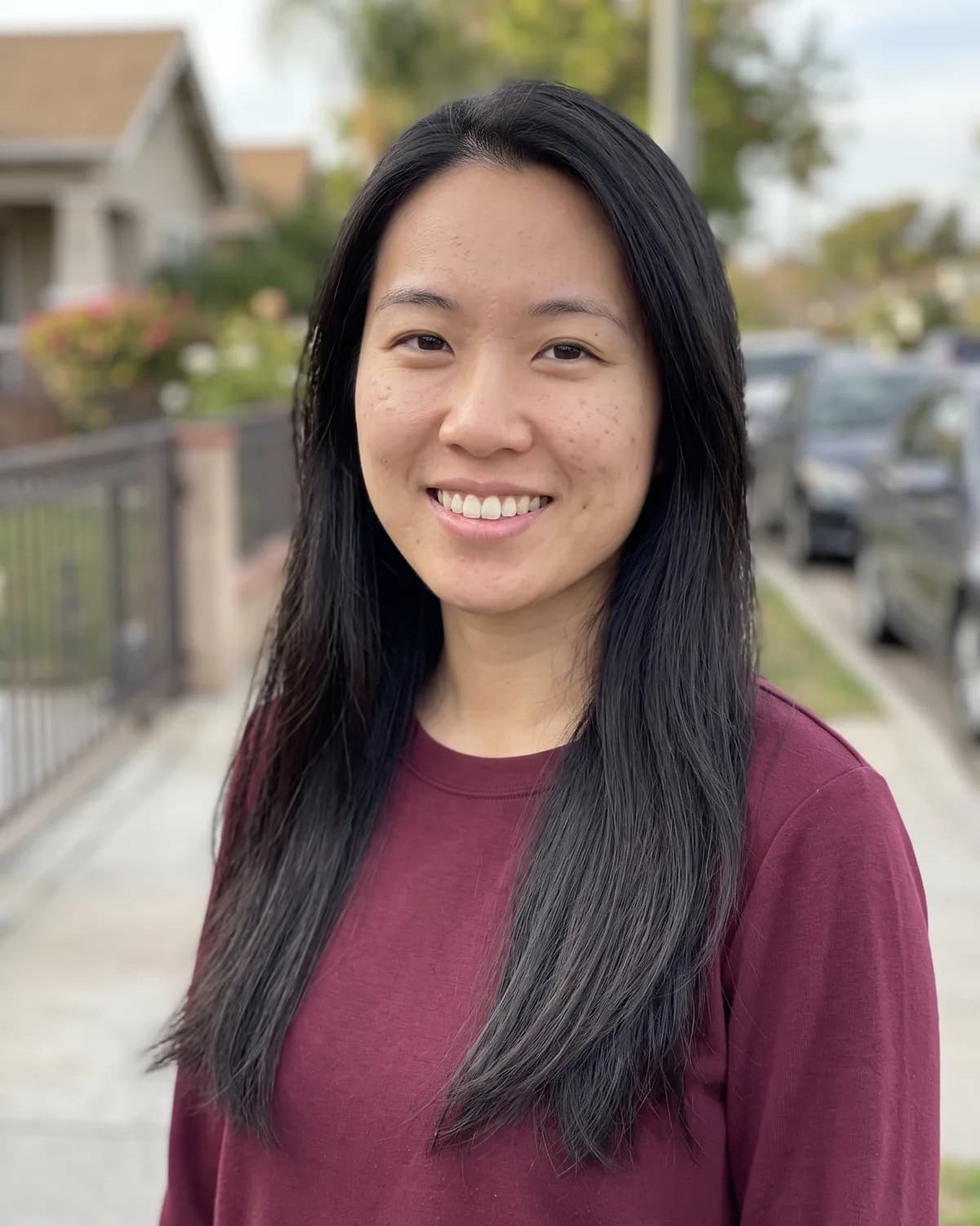 East Asian woman with long straight black hair in a maroon sweatshirt, smiling on a residential street.