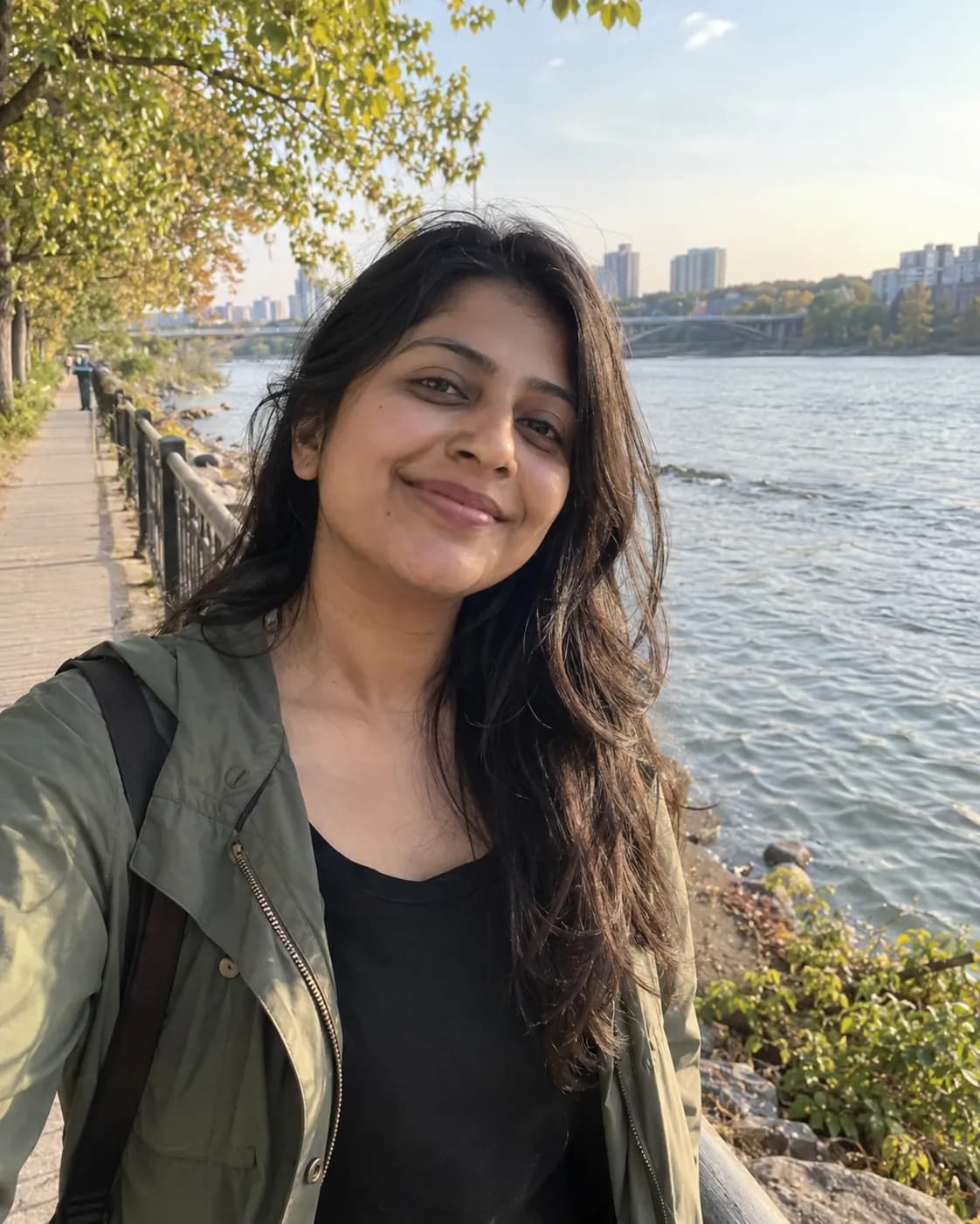 Woman in her late 20s in a sage-green jacket over a black top, on a riverside walkway.