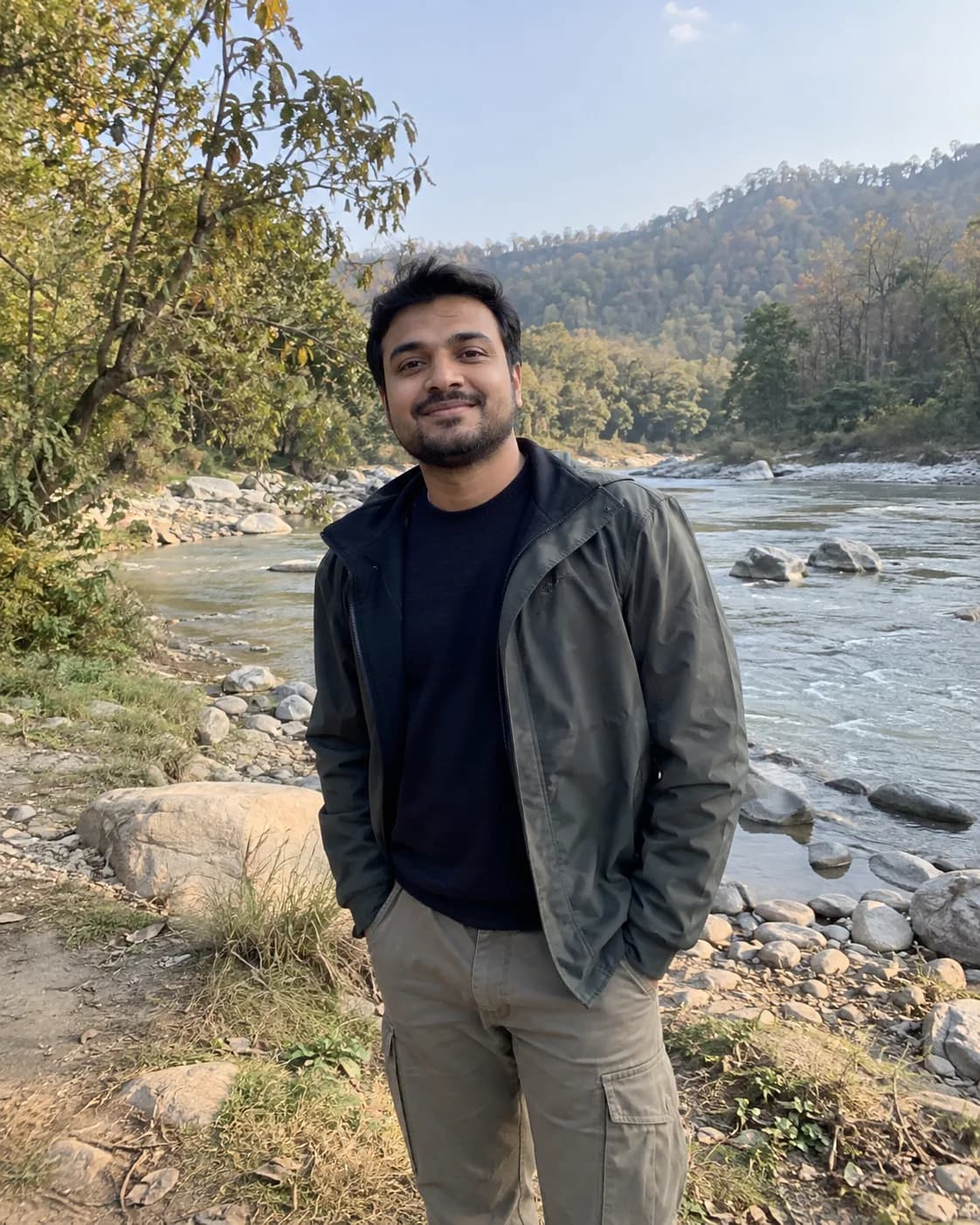 Man with stubble in a dark-green jacket over a black tee, on a rocky riverbank.