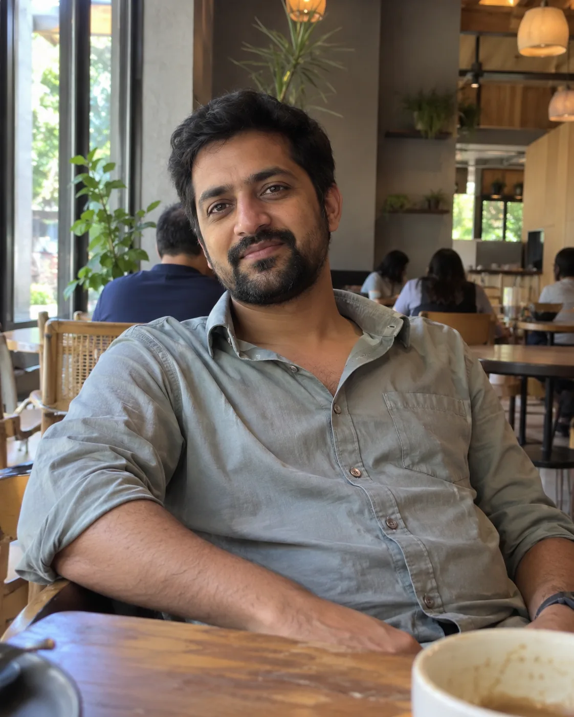 Bearded man in a grey button-up at a cafe table by a window.