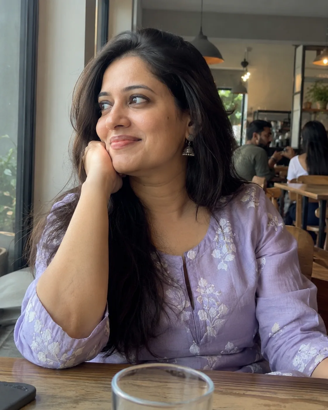 Woman in a lilac embroidered kurti, hand at her chin, at a cafe table.