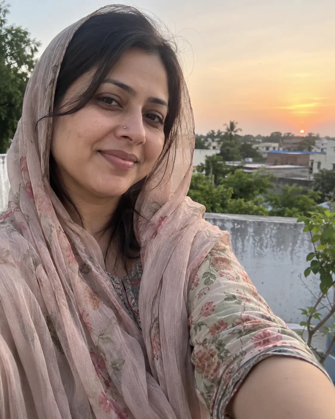 Woman in a pink dupatta and floral kurti, selfie against a sunset sky.