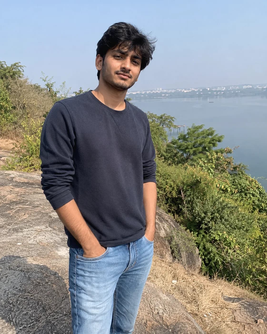 Young man in a navy sweatshirt and light jeans, with greenery and water behind.