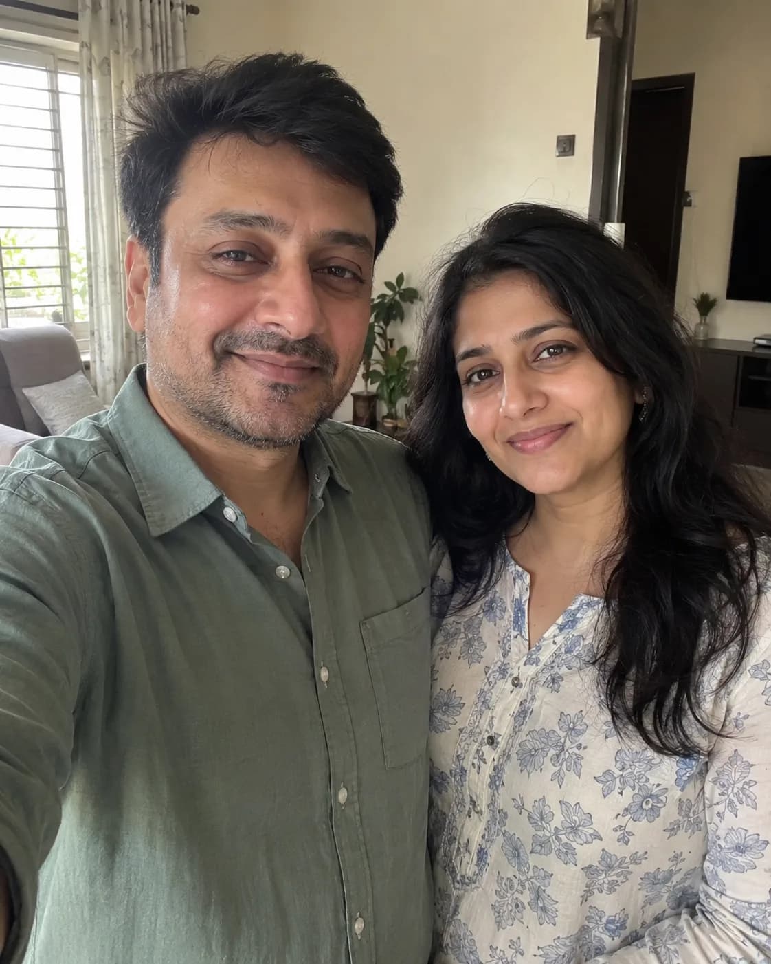 Couple in their 40s in a living room, him in a sage-green shirt and her in a cream and blue floral kurti.