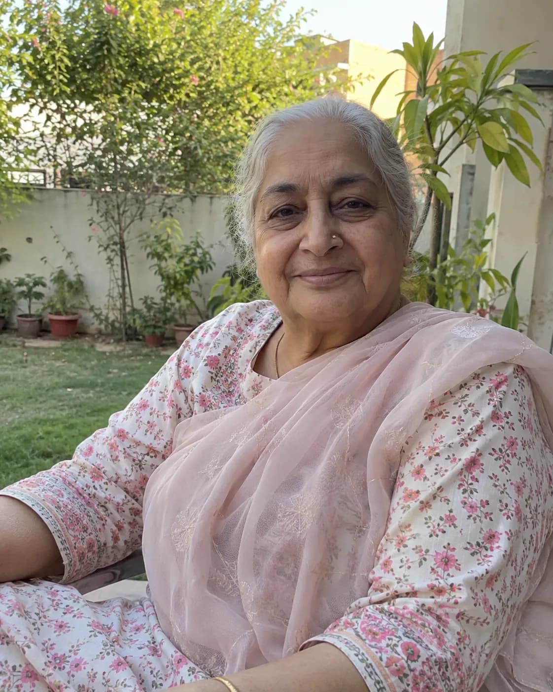 Grandmother about 70 with grey hair in a pink floral sari, in a courtyard garden.